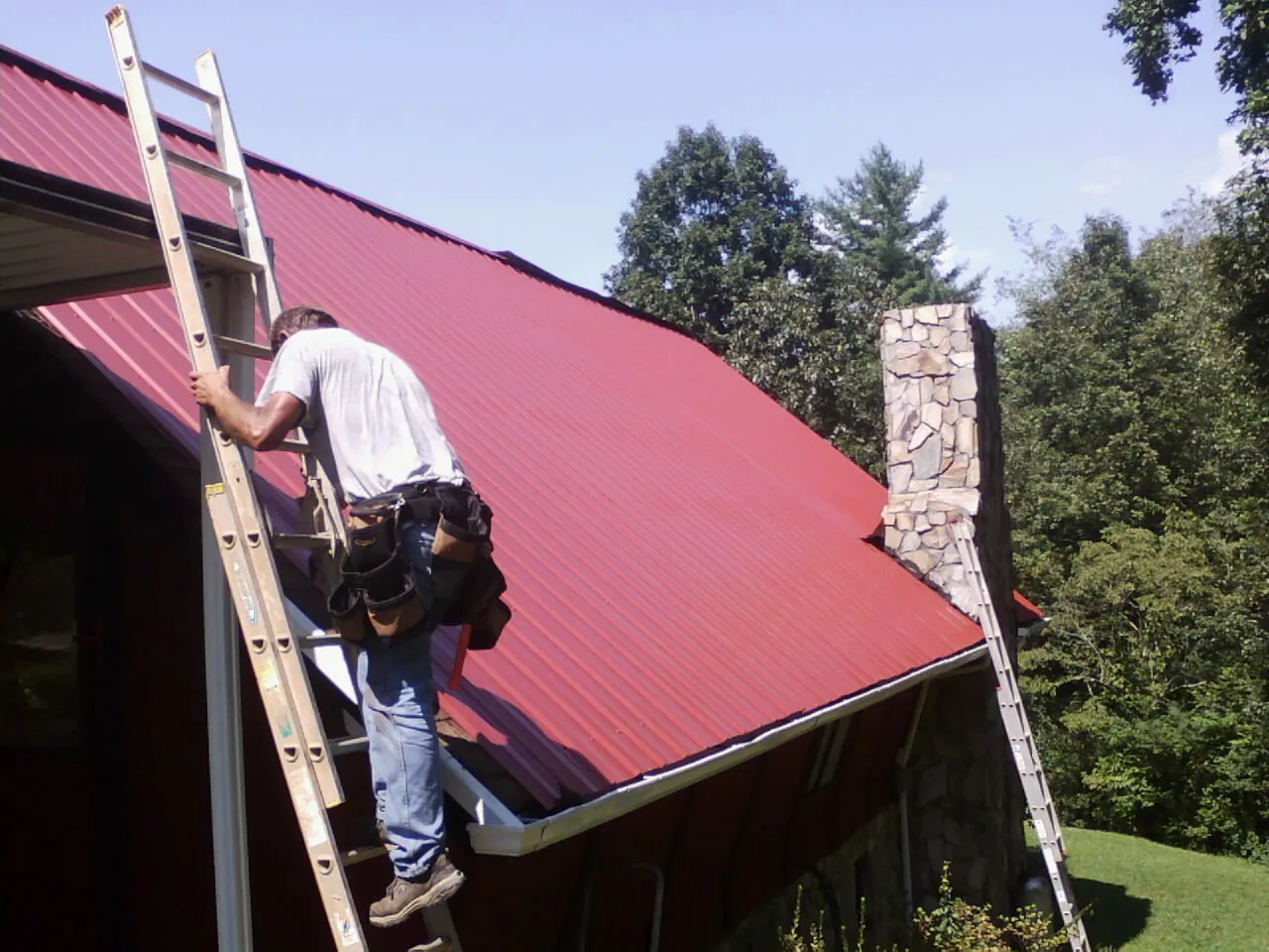 Roofer on a ladder beside a red metal roof and stone chimney — Soffit & Fascia Repair by Roofing Brothers in Tunkhannock